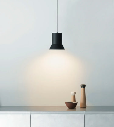 A modern black pendant light with a conical shade hangs over a countertop, casting a warm glow downwards. The scene includes a light gray wall, a gray stone or concrete counter, and two small decorative vases on the right side.