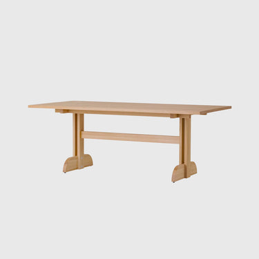 A light natural wood dining table with a rectangular top, a central support beam, and two angled legs ending in rounded feet, shown on a plain light grey background.