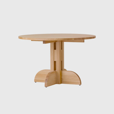 A light natural wood pedestal dining table with a round top and a unique split base, shown against a solid light grey background.