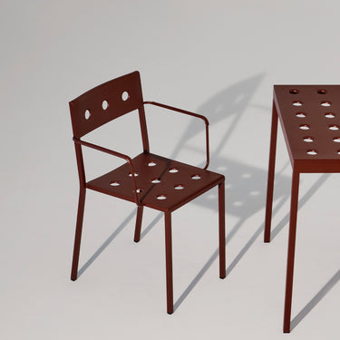 A dark reddish-brown metal chair with armrests and circular cutouts, shown beside a matching table on a light gray studio background.
