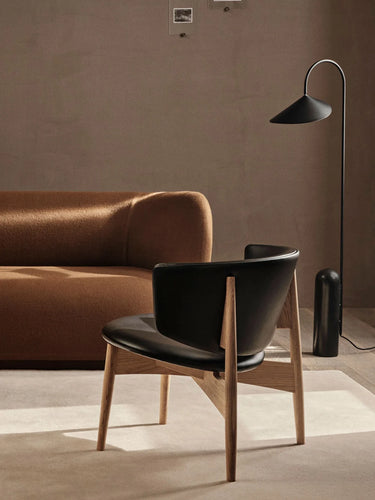 A modern lounge chair with a black leather seat and curved backrest, supported by light oak legs, styled in a minimalist living room next to a brown sofa and a black floor lamp.