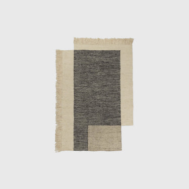 A rectangular woven rug with a geometric pattern, featuring a dark charcoal gray central block, a beige lower-right block, and a cream outer border with fringed edges, shown against a light gray background.