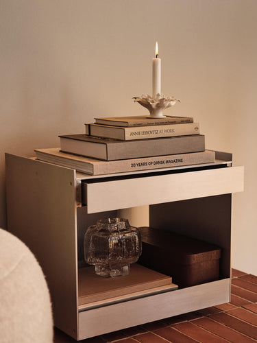 A modern metallic side table with an open shelf and a pulled-out drawer, styled with stacked books, a candle, and a glass vase in a warm indoor setting.