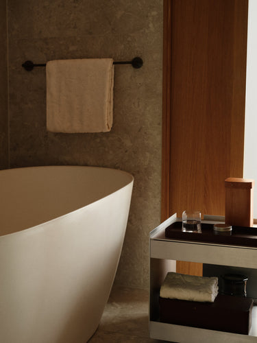 A modern freestanding bathtub in a warm-toned bathroom setting, with a beige towel hanging on a black wall-mounted rack and a small side table holding toiletries beside a wooden door.