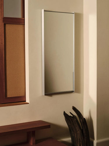 A tall, narrow rectangular wall mirror with a slim silver frame, mounted on a beige wall in a styled interior setting with a wooden bench and decorative trunk.