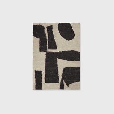 An abstract area rug with a textured, shaggy surface featuring bold black geometric shapes on a warm beige background, shown against a neutral light gray backdrop.