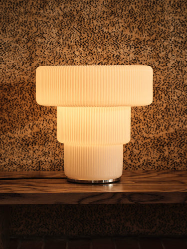 A modern three-tier table lamp with a ribbed, frosted glass shade and a silver metal base, glowing with a warm light against a textured brown and black speckled wall.