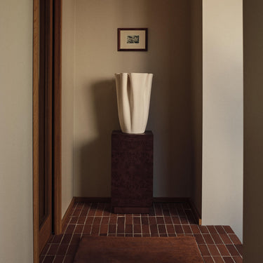 A modern sculptural white ceramic vase displayed on a dark brown textured pedestal in a minimalist hallway with brown tiled flooring and beige walls.