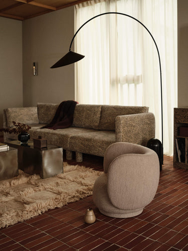 A modern living room scene featuring a textured beige sectional sofa, a matching round armchair, a large black arched floor lamp, and a dark wood coffee table on a shag rug over reddish-brown brick flooring.