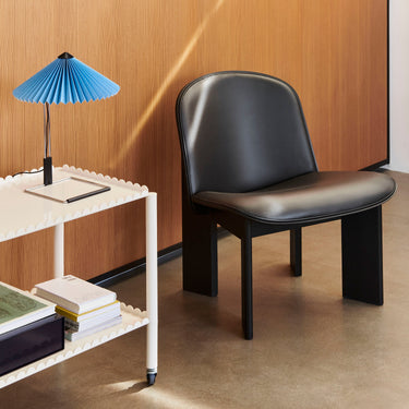 A black upholstered lounge chair with a smooth, streamlined design, positioned next to a white side table with a blue lamp on it, against an interior background.