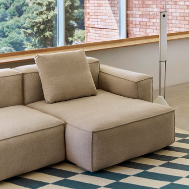 A modern modular sectional sofa in a light beige linen fabric, featuring a low-profile chaise lounge section with a matching throw pillow, set in a contemporary room with a checkered rug and large windows.