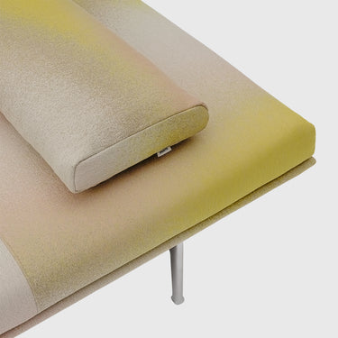 A close-up of a modern upholstered sofa or daybed with a matching rectangular pillow, showing a soft gradient fabric that shifts from pale beige to muted yellow-green and warm yellow. A slim metal leg is visible against a clean light gray background.