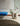 A modern beige ceramic U-shaped planter vase filled with wildflowers, placed on a light tiled floor in a minimalist interior setting with a blue bench and a brown textured rug in the foreground.