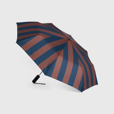 A closed folding umbrella with alternating wide stripes in navy blue and muted terracotta brown, shown open against a light gray studio background.