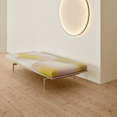 A modern low-profile daybed with a white and yellow abstract patterned cushion, supported by slim metal legs, set in a minimalist room with light wood flooring and a warm glowing circular wall light.