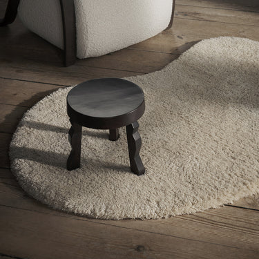 A small round dark wood stool with turned legs sits on a cream shag rug over a rustic wooden floor, with part of a white upholstered chair visible in the upper left.