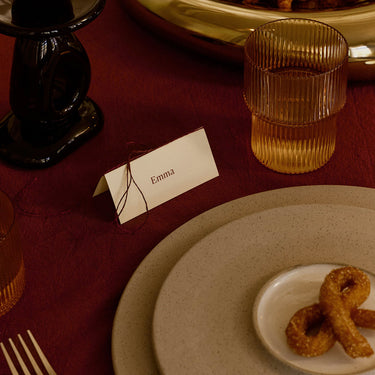 A close-up of a styled dining table setting with a cream paper place card standing on a dark red tablecloth, amber ribbed glass tumblers, layered beige speckled plates, and a small dish of fried appetizers.
