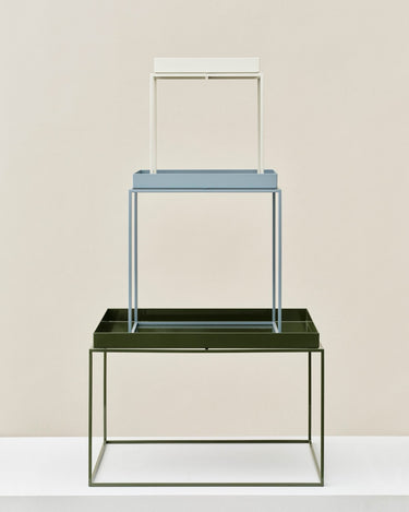 A minimalist three-tier display stand or shelving unit with a dark olive green base, a light blue middle tier, and a white top frame, set against a warm beige background.