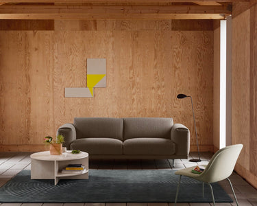 A modern living room featuring a taupe fabric sofa, a white round coffee table, a cream molded chair, a dark blue patterned rug, and a light wood wall with a minimalist yellow and white geometric wall art piece.