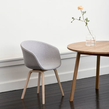 A modern chair with a gray upholstered seat and a beige oak frame, with four legs, placed next to a round wooden table with a vase of flowers on it, set against a white wall on a dark floor.