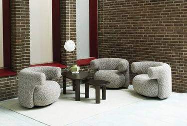Three rounded boucle armchairs in a light gray textured fabric arranged around a small dark wood coffee table on a white rug, set against a brick wall and window.