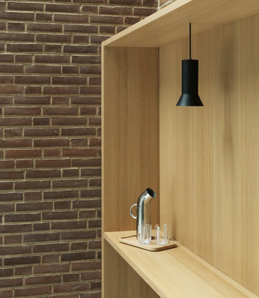 A modern black pendant light hanging above a light wood shelf with a metal water pitcher and two small glasses, set against a brick wall background.
