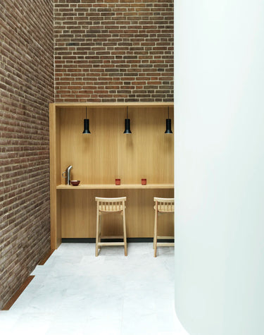 A minimalist interior scene featuring a light wood bar counter and two matching wooden stools in front of a brick wall, with pendant lights above and a white curved wall on the right.