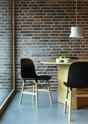 A modern dining nook with a round light wood table, two black chairs with light wood legs, set against a rustic red-brown brick wall and a large window.