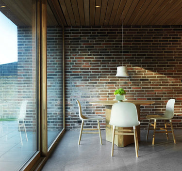 A modern dining area with a round wooden table, three white chairs with light wood legs, a hanging pendant light, and a brick wall backdrop next to large glass windows.
