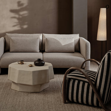 A modern living room scene featuring a light beige bouclé sofa with two matching back cushions, a low octagonal cream coffee table, and a black-and-cream striped armchair with curved wooden arms. A tall floor lamp stands against a dark brown wall with soft shadows cast across it.