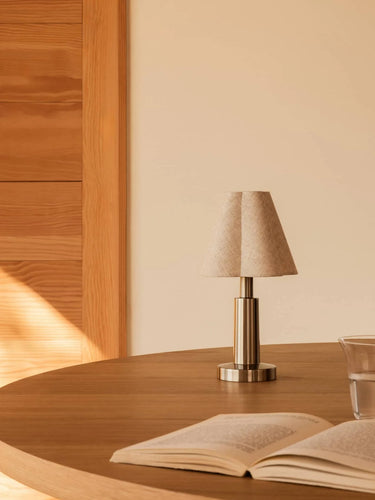 Table lamp on a wooden table with an open book and a glass of water, against a beige wall.