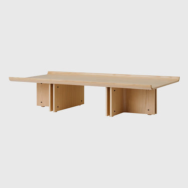 A minimalist light wood table with a rectangular top and three geometric block-style legs, shown against a solid light grey background.