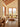 A minimalist dining area featuring a round wooden table and two matching chairs, positioned in front of large windows that offer a view of rolling green hills. The room has warm beige walls, exposed wooden ceiling beams, and a matching wooden floor.