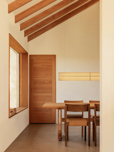 A minimalist dining area featuring a wooden table and chairs, a warm wood door, and a slanted ceiling with exposed wooden beams under a cream-colored wall.