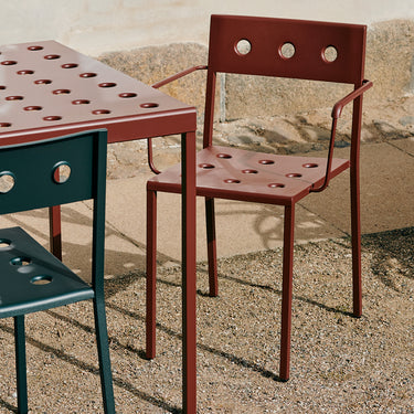 A red metal outdoor dining chair with a perforated seat and back, shown next to a matching table and another dark green chair on a gravel patio.