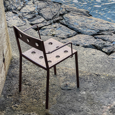 A dark brown metal chair with a perforated seat and back, positioned on a rocky, concrete surface next to water.