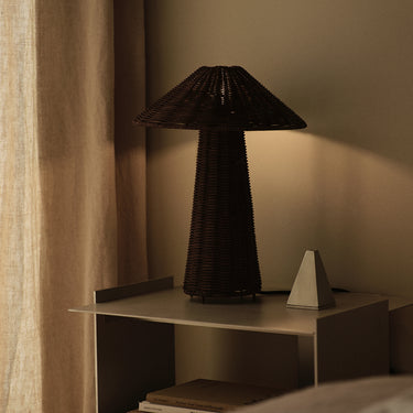 A dark brown woven table lamp with a conical shade and textured base, illuminated on a light gray side table next to a small white geometric object in a warm, dimly lit room.