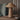 A rustic wicker table lamp with a conical shade and textured woven base, styled on a wooden table next to a green armchair and a dark ceramic vase.