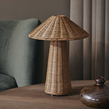A rustic wicker table lamp with a conical shade and textured woven base, styled on a wooden table next to a green armchair and a dark ceramic vase.