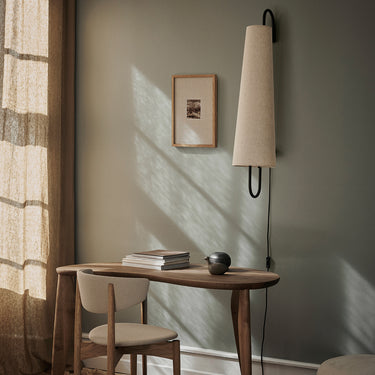 A modern wall sconce with a tapered beige fabric shade and black metal U-shaped arm, mounted on a sage green wall in a styled home office setting with a wooden desk, chair, and warm sunlight.
