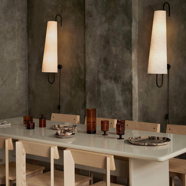 A dining room scene with a long white table, light wood chairs, amber glassware, and two tall wall sconces mounted on textured dark grey walls.
