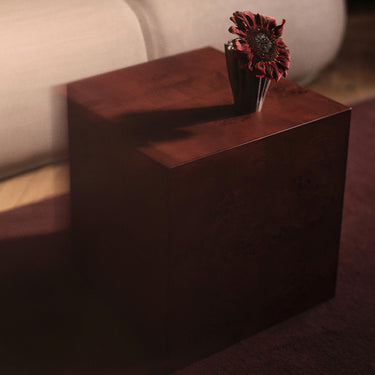 A dark reddish-brown wooden cube side table or end table with a single dried red flower in a dark vase on top, set in a dimly lit interior next to a light-colored sofa.