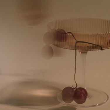 A clear ribbed glass coupe-style coupe glass with a stem, shown on a warm beige background with grapes hanging from the rim and base.