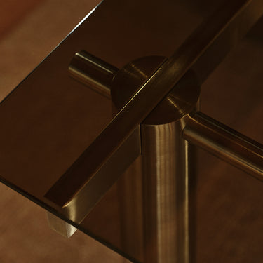 Close-up detail of a modern table with a brushed brass or bronze metal frame and glass top, showing the corner joint and leg construction.