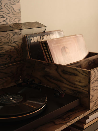 A wooden record player with a transparent dust cover and vinyl records inside a matching wooden storage box, set against a warm beige background.