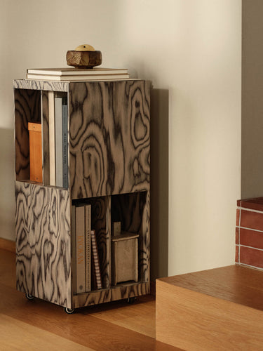 A two-tier rolling bookcase with a distinctive wavy wood grain pattern in gray and black, styled with books and a small decorative bowl on top, set in a minimalist room corner.