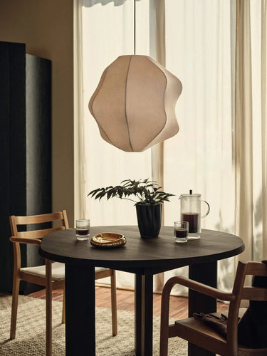 A modern pendant light with an organic, wavy paper-shield design hangs over a round dark wood dining table in a warm, sunlit room with wooden chairs and sheer curtains.
