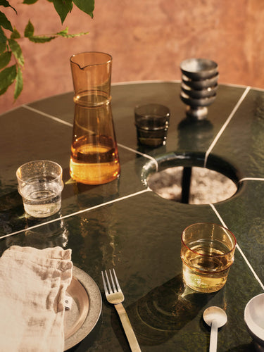 Table setting with a carafe, glasses, and cutlery on a reflective surface.