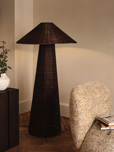 A dark brown woven floor lamp with a tapered conical shade and textured base, styled in a warm, neutral-toned room next to a textured armchair and a side table.