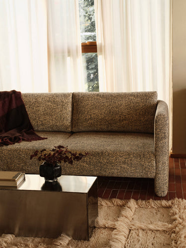 A two-seater upholstered sofa in a textured beige and brown bouclé fabric, styled in a warm living room setting with a dark red throw blanket, a reflective metal coffee table, and a shag rug.